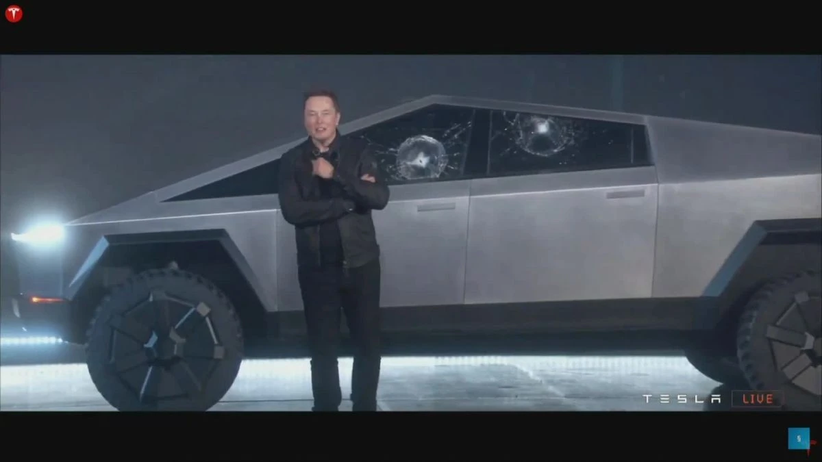 Screenshot of Elan Musk standing in front of a Tesla with broken windows. 